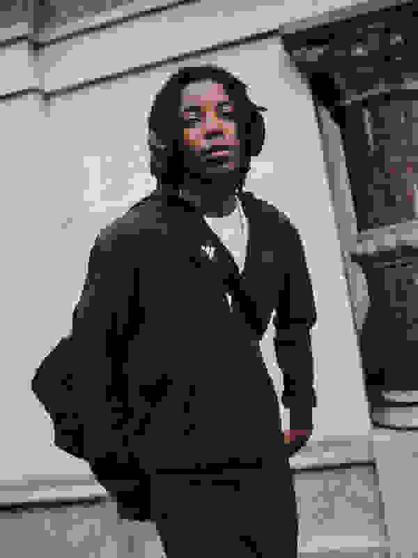 Siyah kapüşonlu bir ceket giyen saçlarıyla genç bir adam taş bir binanın önünde duruyor, onun ifadesi düşünceli.