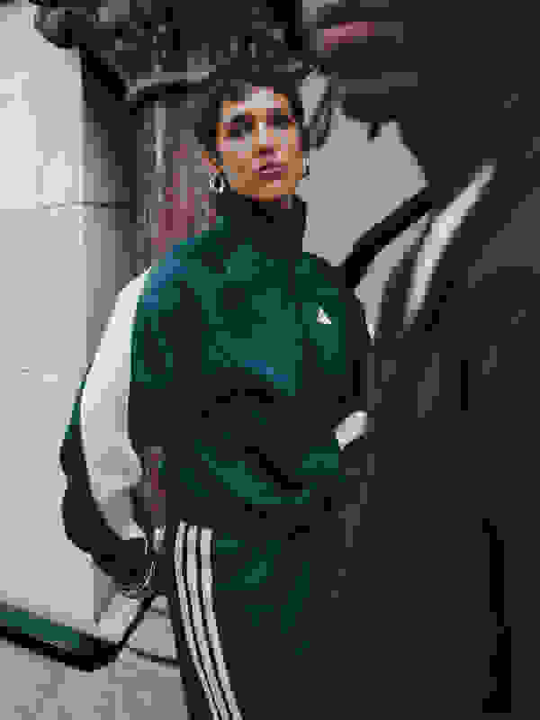 A person wearing a green Adidas tracksuit stands in front of a brick wall, with another person visible in the background.