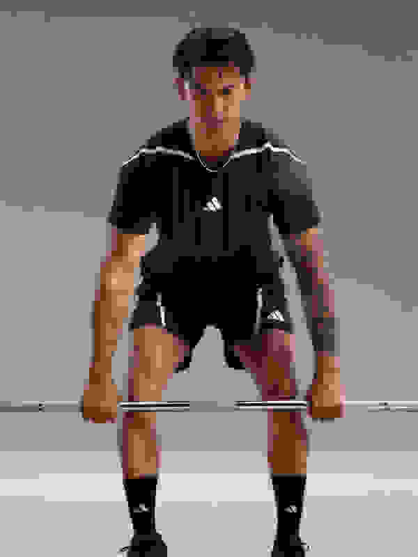 A muscular man in a black athletic shirt and shorts is crouched down, gripping a barbell in front of a plain gray background.