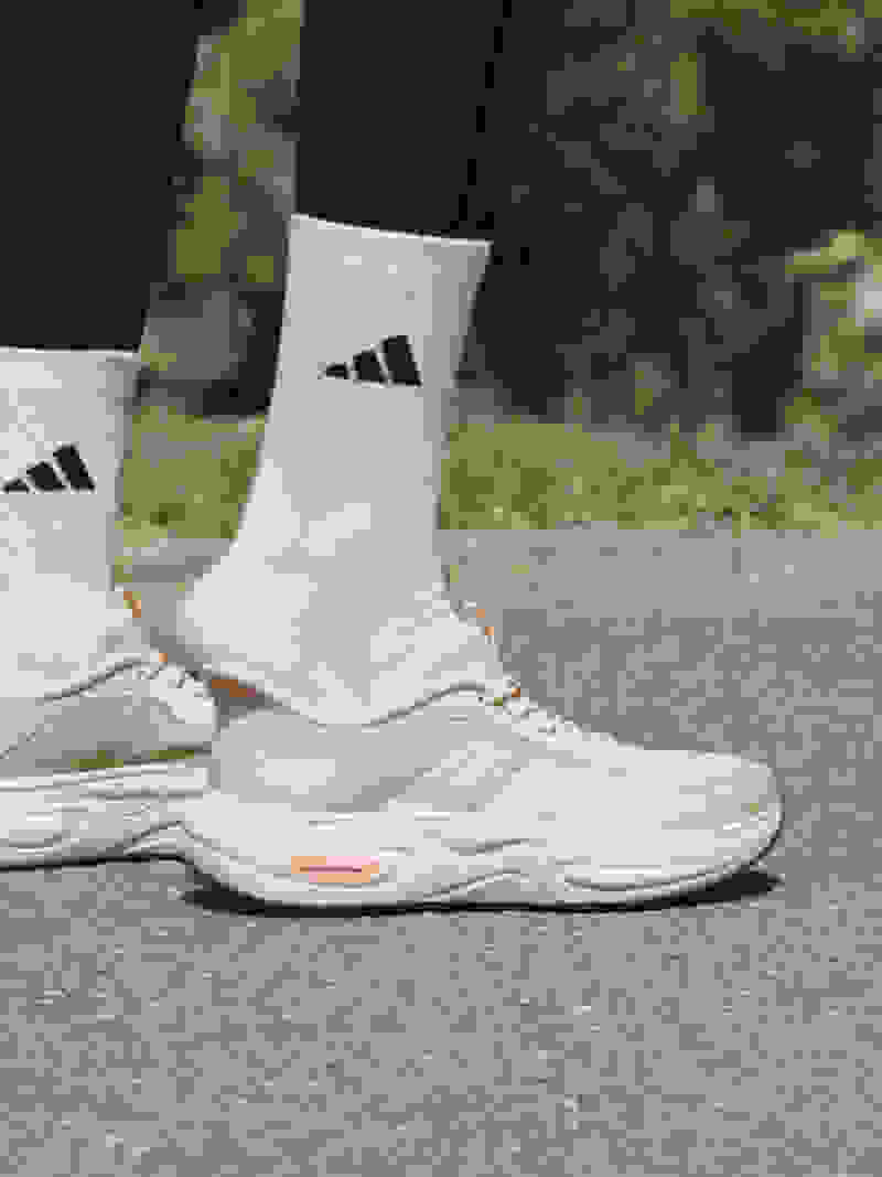 The image shows a pair of white Adidas sneakers with black stripes on a person's feet, standing on a paved road with a lush green background.