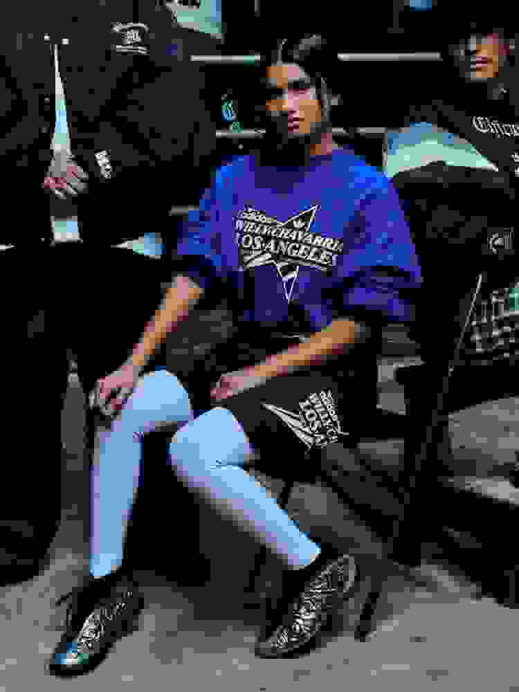 Close-up of a woman wearing a blue adidas x Willy Chavarria 'Los Angeles' top with a large star graphic and Trefoil logo on the front. She stands with her arm resting on the shoulder of a person wearing a black “Chicano” adidas Originals x Willy Chavarria top.