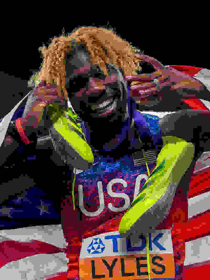 A smiling athlete wearing a USA jersey and holding an American flag, with a dark background.