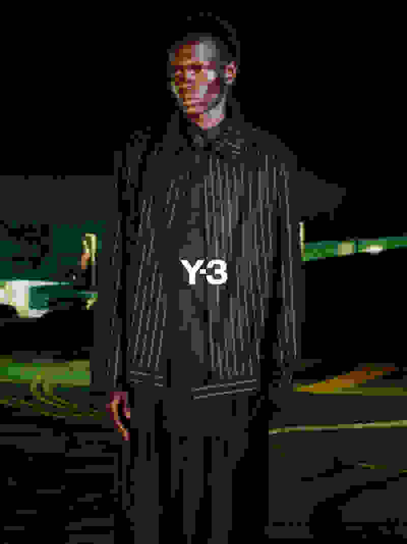 A person wearing a Y-3 branded jacket stands in a dimly lit outdoor setting, with a blurred background suggesting a car park or similar urban environment.
