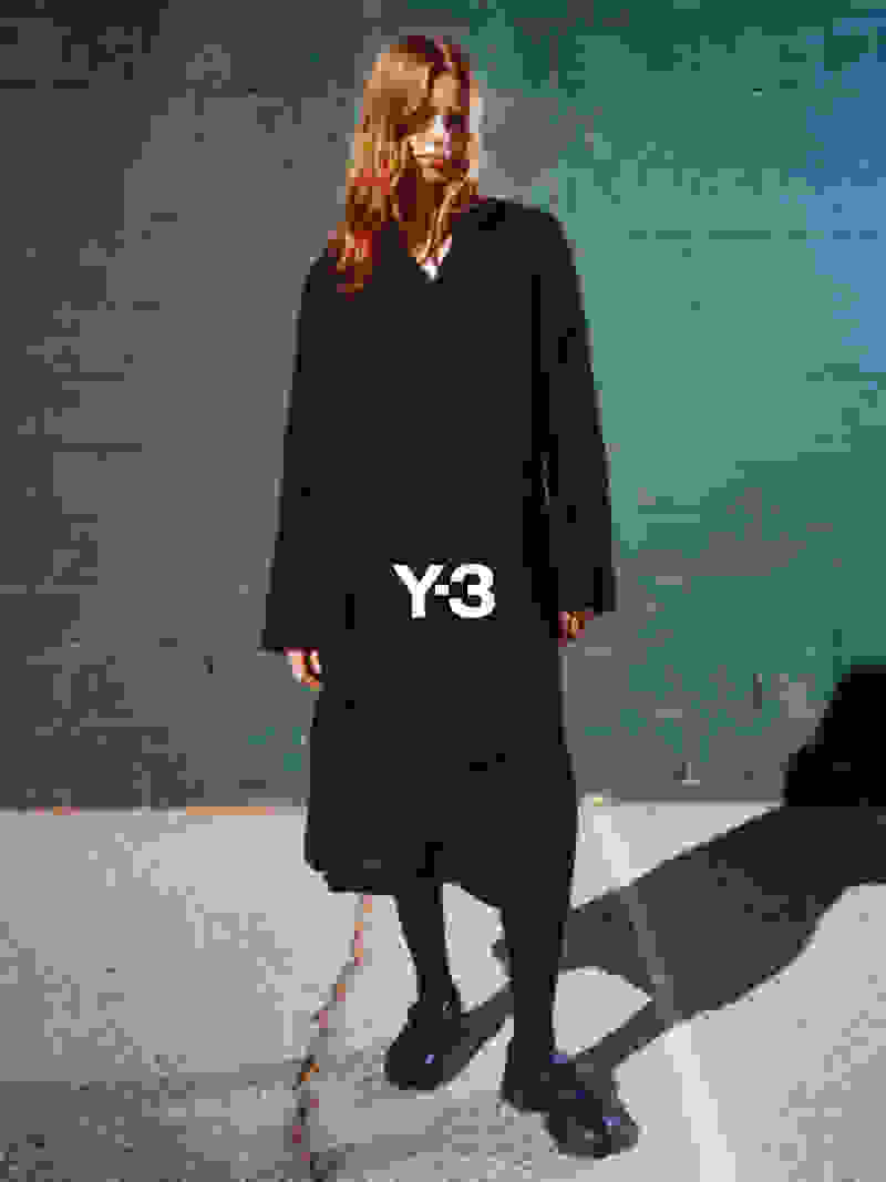 A young woman with long, reddish-brown hair stands in front of a concrete wall, wearing a black Y-3 jacket and matching trousers, and black boots.