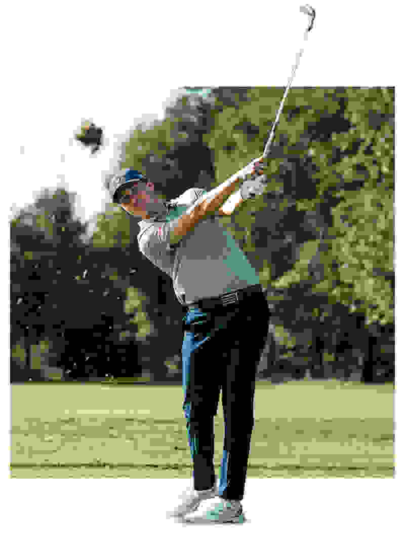 A golfer in a light-colored shirt and dark pants is swinging a golf club on a grassy golf course surrounded by trees.