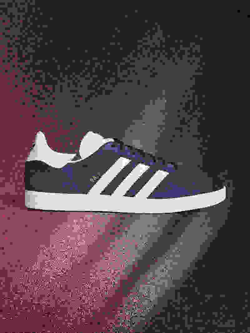A navy blue and white Adidas Gazelle sneaker is displayed on a red and black textured background.