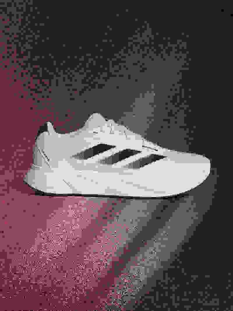 A white and black athletic shoe is prominently displayed against a dark, textured background with a reddish-pink gradient.