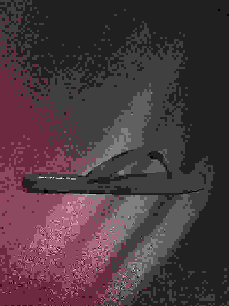 A black flip-flop sandal is shown against a dark, textured background with a reddish hue.