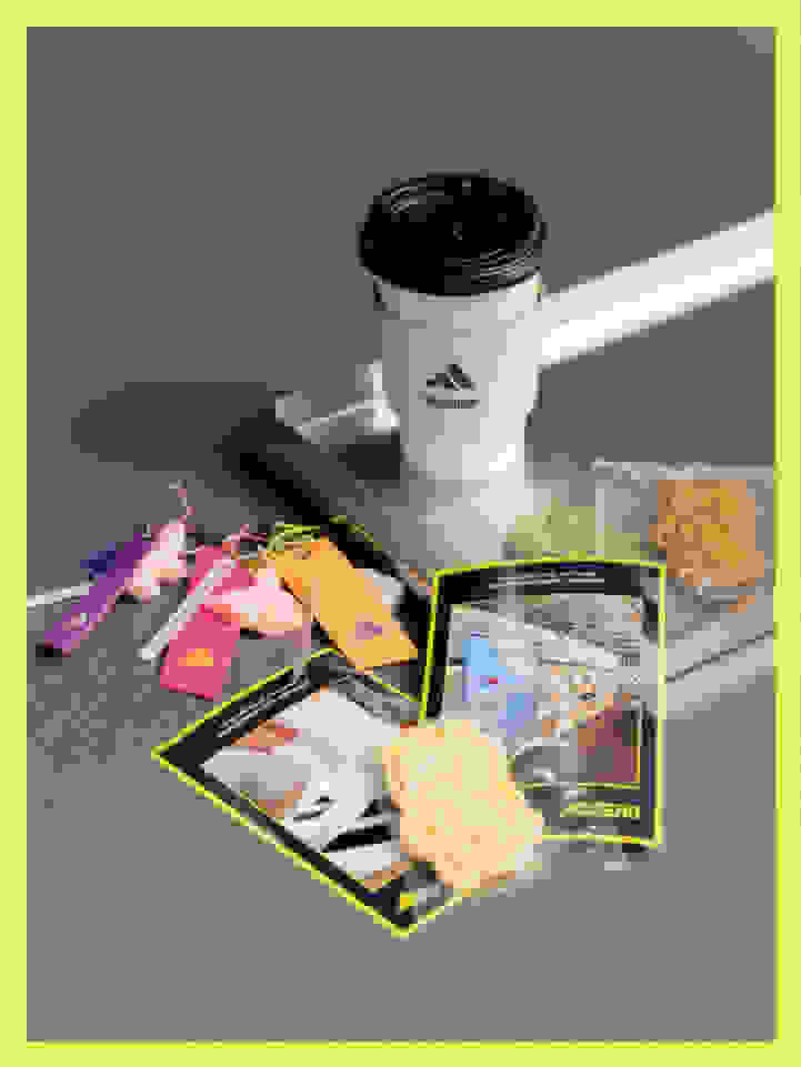 The image shows an assortment of items, including a white coffee cup with a black lid, a tablet device displaying an image, and various colorful objects such as stickers and snacks scattered on a gray surface.