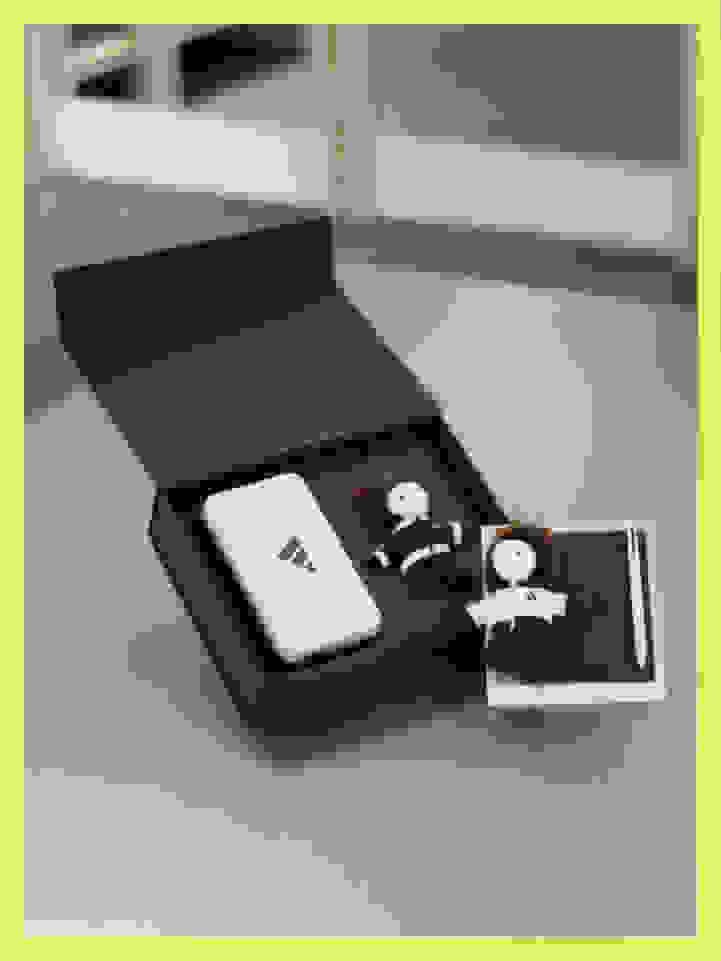 A black box-like container with a white device and two small figurines inside, resting on a light-colored surface.