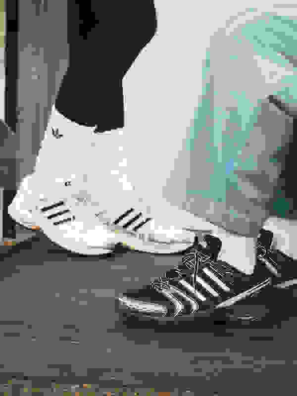 The image shows a pair of white and black athletic shoes on a wooden surface, with a person's legs and clothing visible in the background.