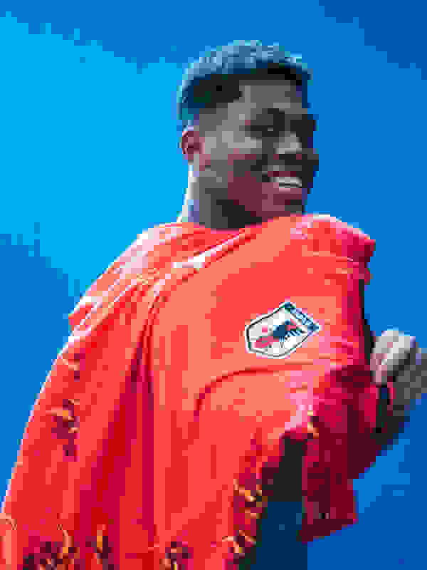 A smiling young man wearing a bright red jacket stands against a vibrant blue background.