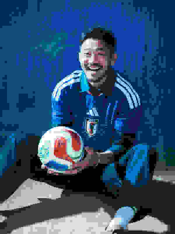 A smiling man in a blue and white sports jersey is sitting on the ground, holding a soccer ball.