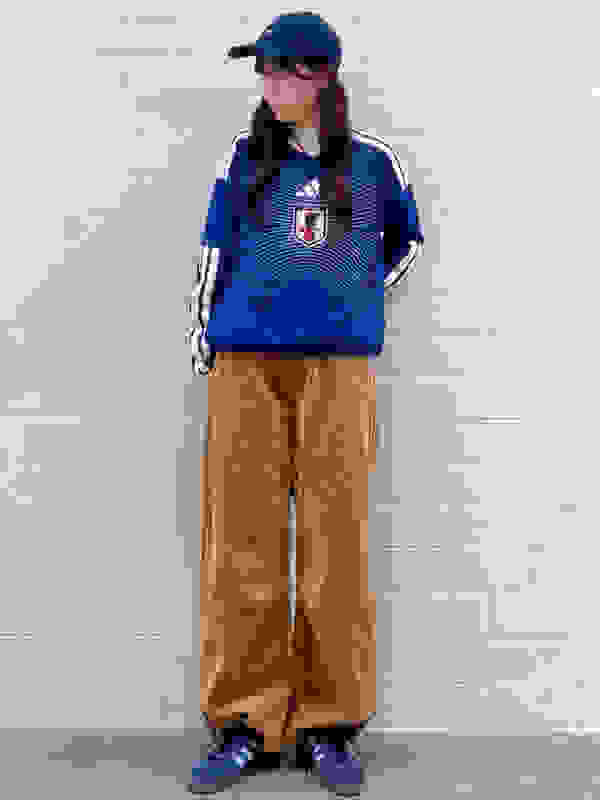 A young woman wearing a blue and white striped jersey, blue baseball cap, and brown corduroy pants stands in front of a white brick wall.