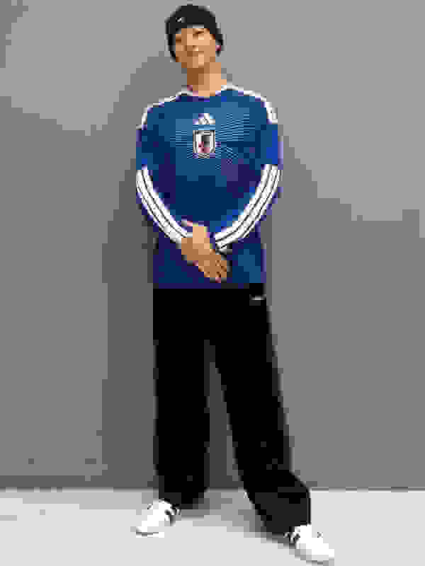 A person wearing a blue and white striped jersey, black pants, and a black cap is standing in front of a gray wall.