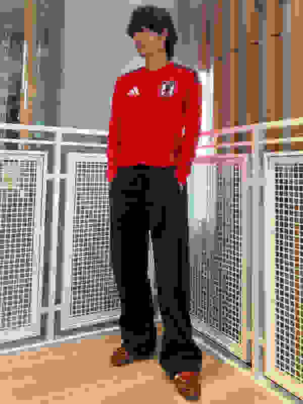 A person wearing a red jersey and black pants stands in a hallway with a metal grate in the background.
