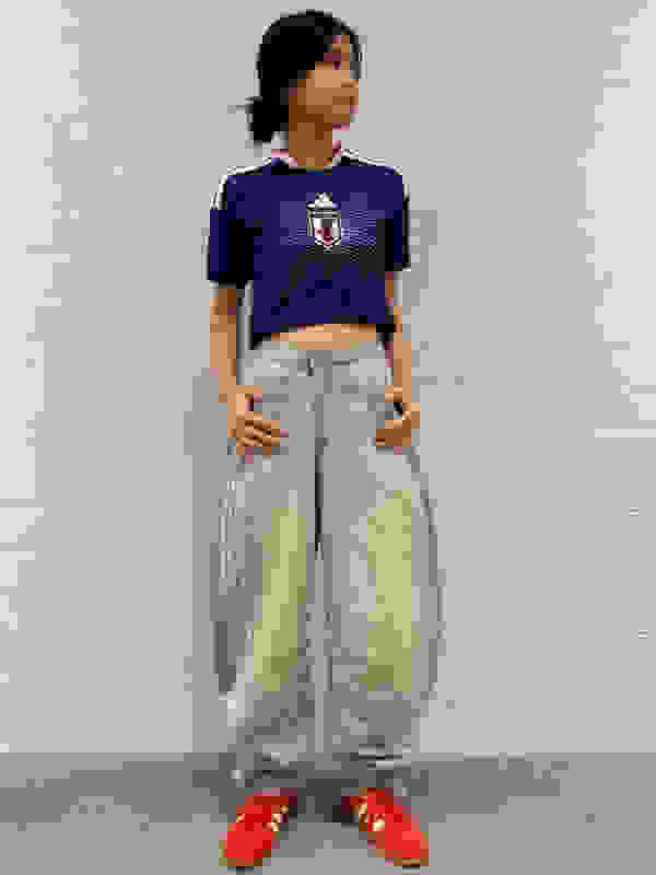 A young person wearing a navy blue sports jersey and loose-fitting gray pants stands in front of a plain white wall.