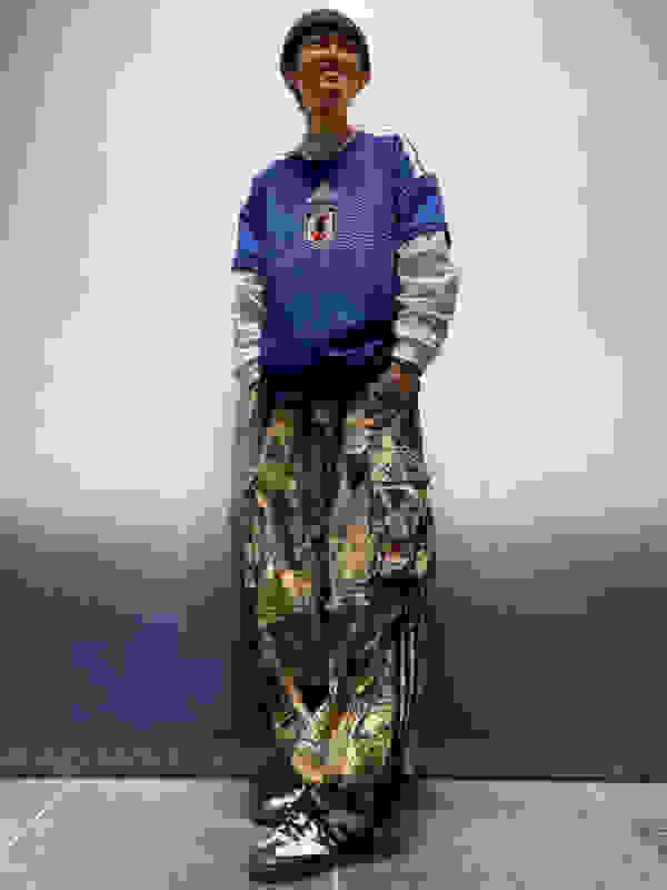 A young person wearing a blue and gray sports jersey, camouflage pants, and black sneakers stands in a plain indoor setting with a blue wall in the background.