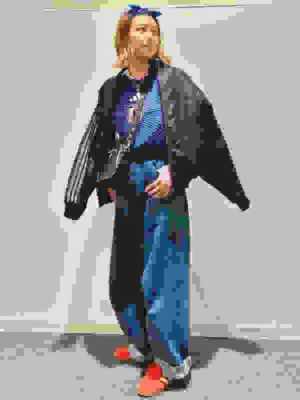 A young woman wearing a black leather jacket, striped top, and loose-fitting jeans stands in a plain, white-walled room.