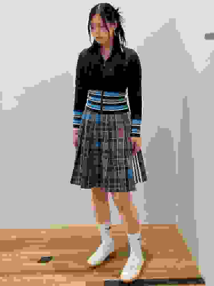 A young person wearing a brown top and a plaid skirt stands on a wooden floor in a plain room.