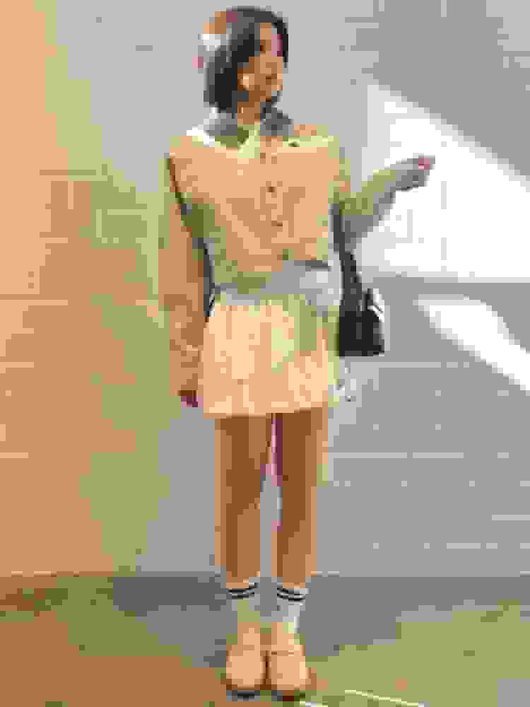 The image shows a young woman wearing a beige jacket and a short skirt, standing in front of a plain white wall.