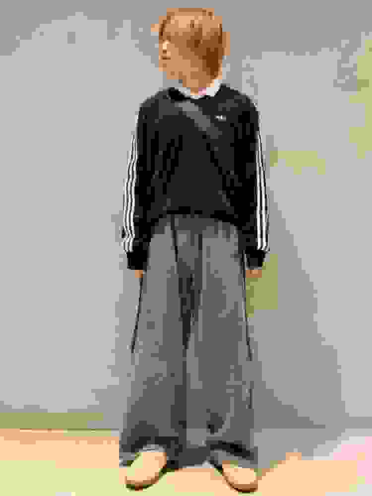 A young person wearing a black top with white stripes and loose gray pants, standing in a plain, light-colored room.