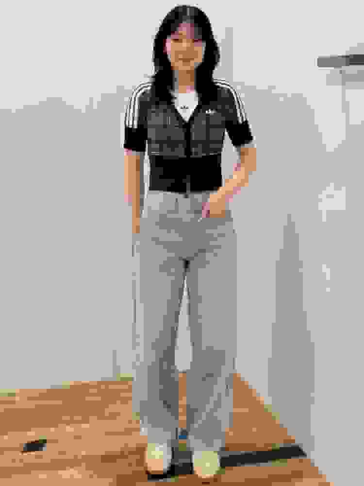 A young woman with dark hair stands in a room, wearing a plaid shirt and light-colored jeans, with her hands in her pockets.