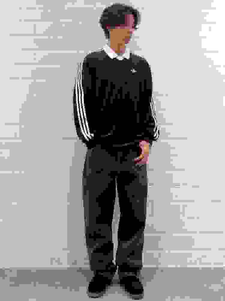 A young person wearing a black long-sleeved shirt with white stripes and dark pants, standing in front of a white brick wall.