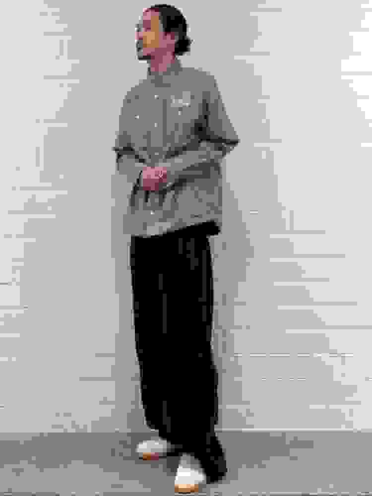 A person wearing a gray button-up shirt and black pants stands in front of a white brick wall.