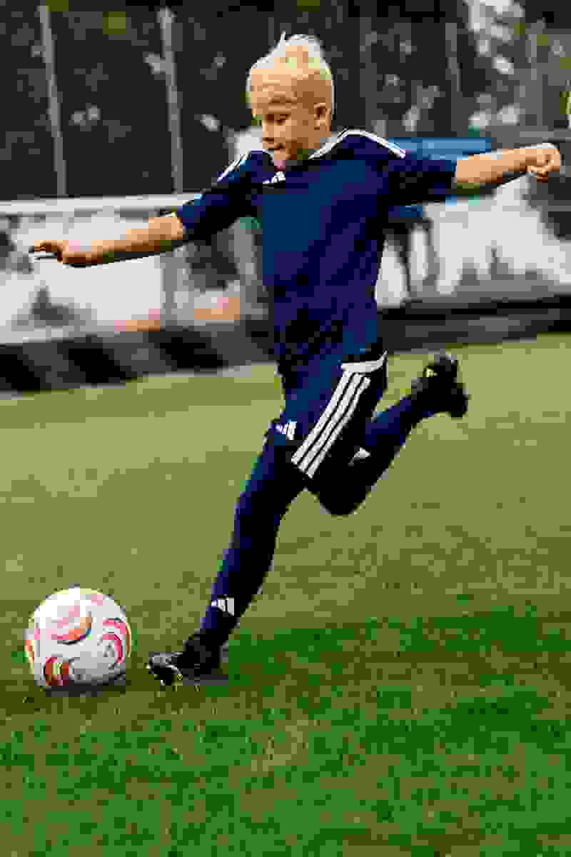 A young boy in a navy blue sports uniform is kicking a soccer ball on a grassy field, with trees and buildings visible in the background.