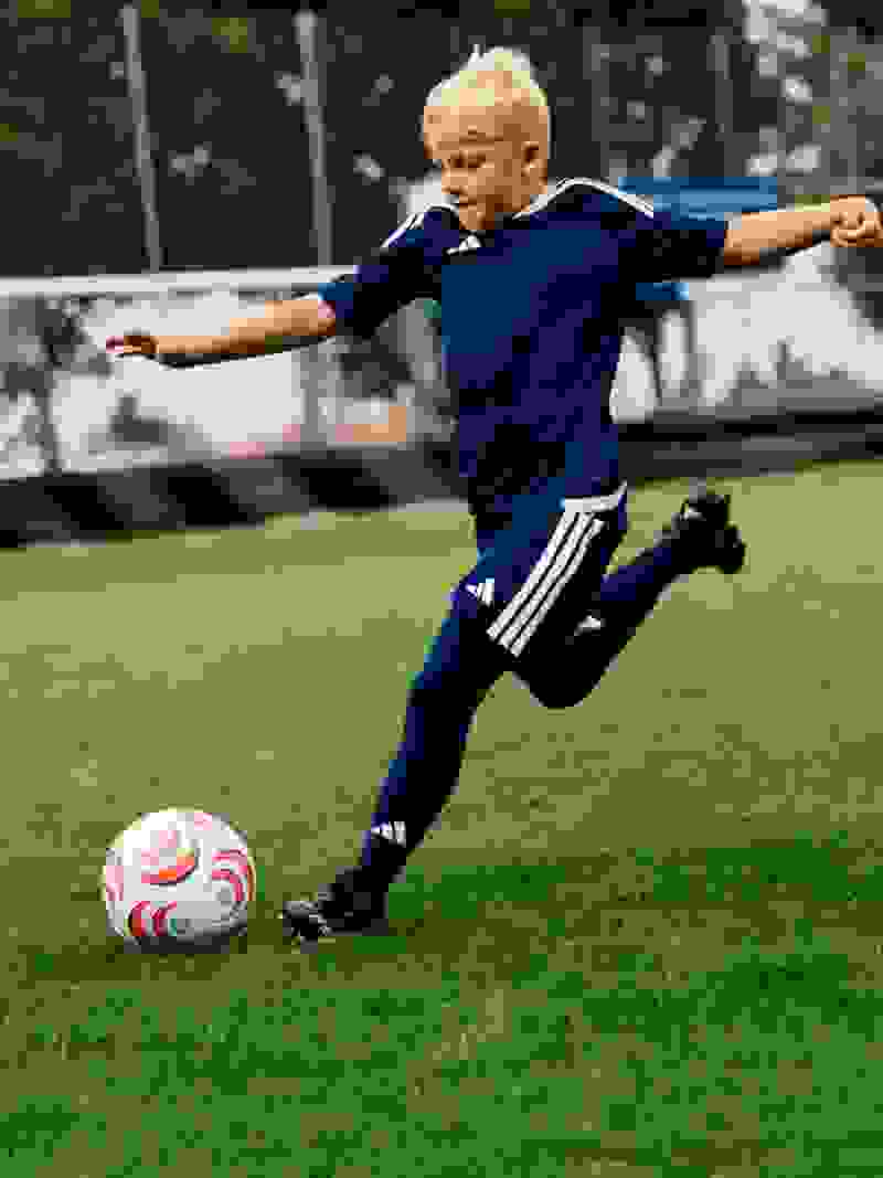 A young boy in a navy blue sports uniform is kicking a soccer ball on a grassy field, with trees and buildings visible in the background.