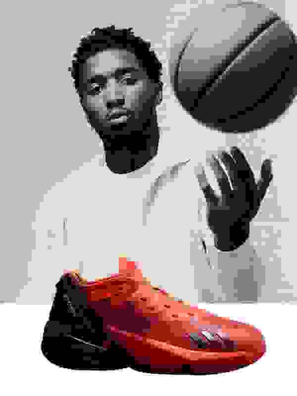 A young man with a beard and curly hair stands in a white room, holding a basketball and wearing a pair of red and black athletic shoes.