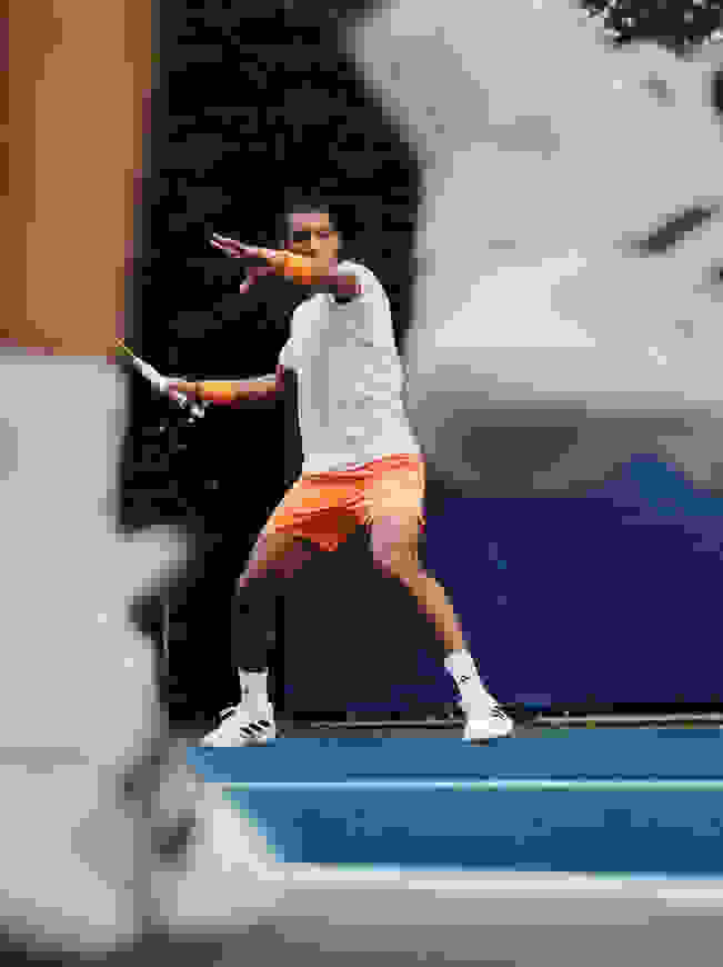 A person in white and orange sportswear is playing tennis on a blue tennis court surrounded by trees.