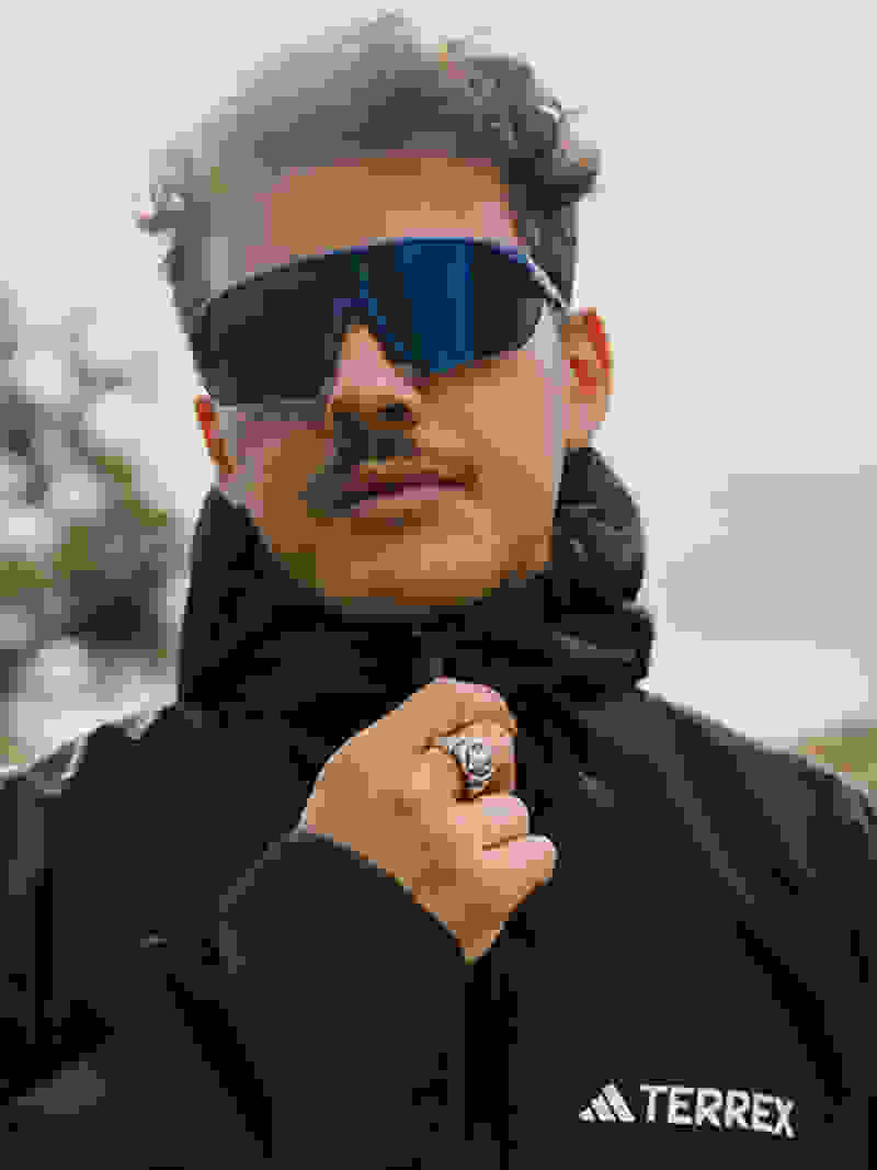 A person wearing a black jacket and sunglasses stands in an outdoor setting with a cloudy sky in the background.