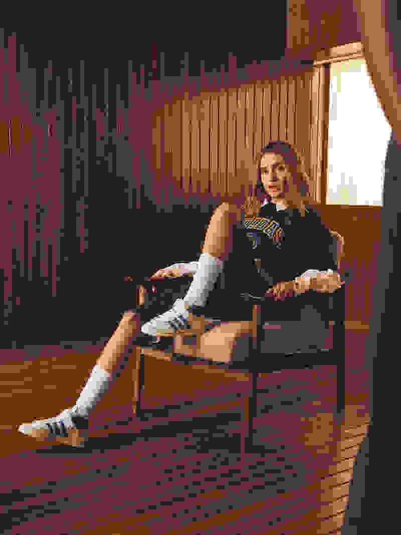 A young woman in casual athletic attire sits comfortably on a wooden chair against a backdrop of wooden paneling.