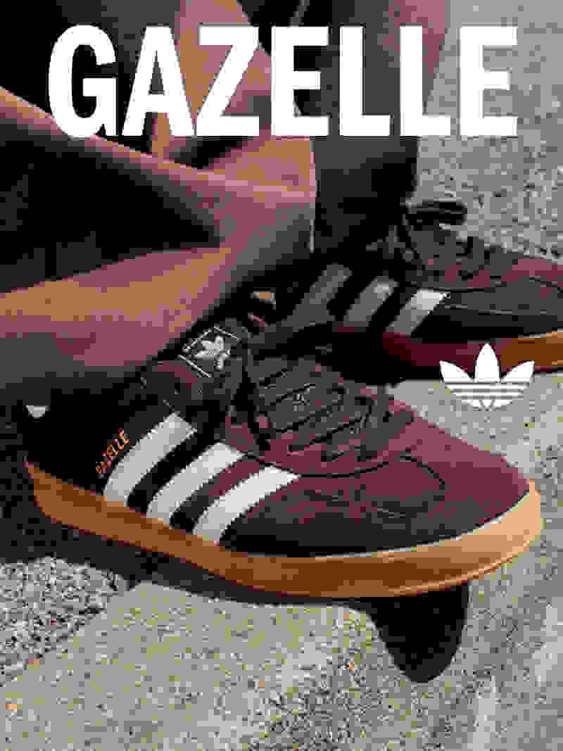 The image shows a pair of Adidas Gazelle sneakers in a burgundy color, with the Adidas logo prominently displayed. The sneakers are resting on a concrete surface, with a blurred background suggesting an urban setting.