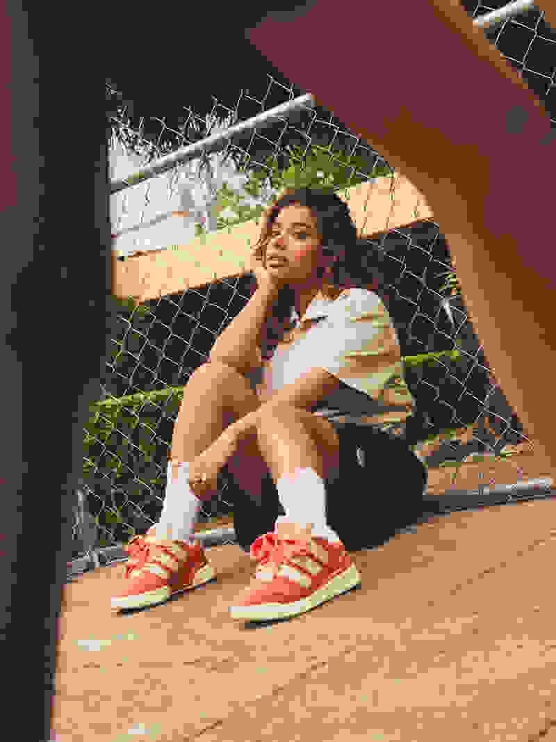 A person with curly hair sits on the ground, wearing a light-colored shirt and red sneakers, against a backdrop of a wire fence and greenery.