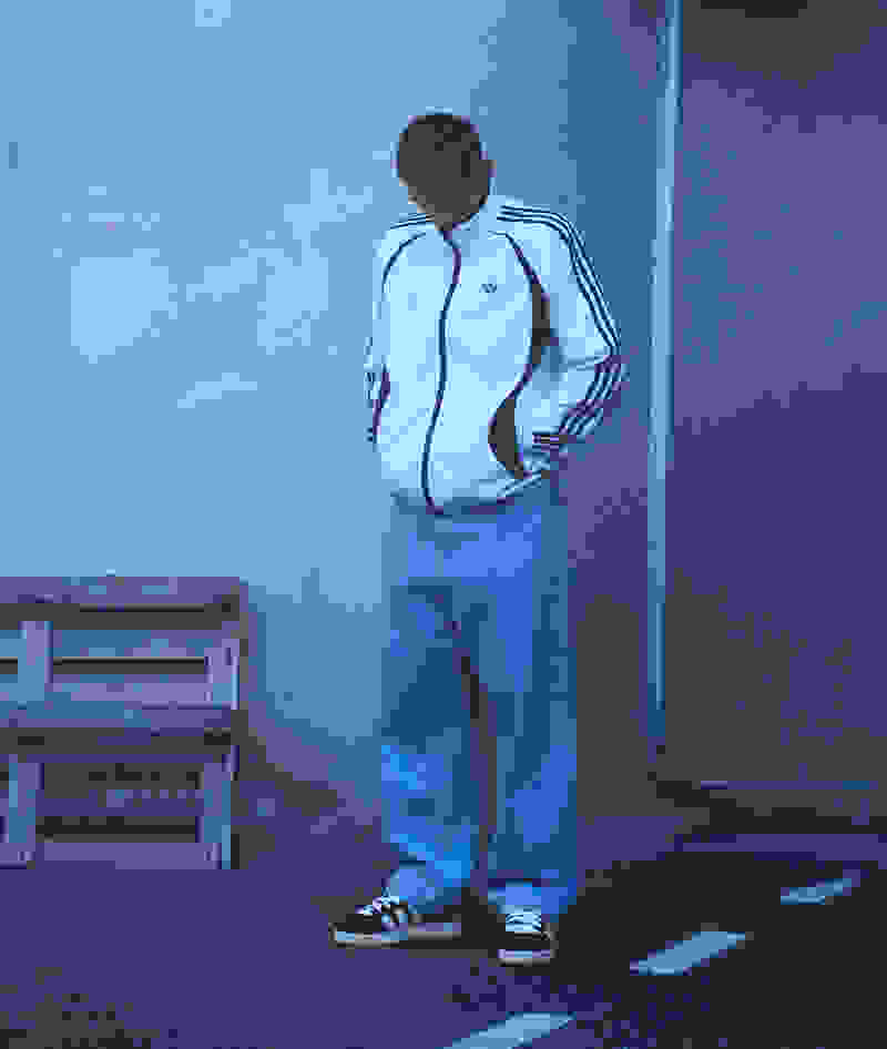 A person wearing a white jacket with stripes and blue pants stands in front of a gray wall, with a wooden crate visible in the background.