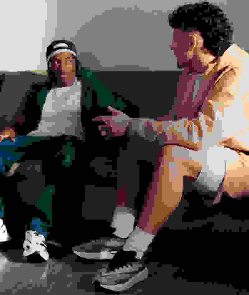Two individuals, one wearing a green jacket and the other in a beige outfit, are seated on a couch engaged in conversation.