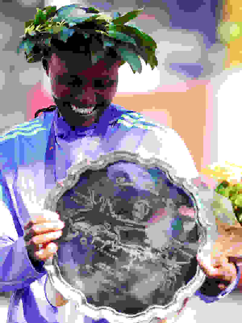 A smiling person wearing a blue and white striped shirt and a wreath of leaves on their head, holding a decorative plate with a celestial design.