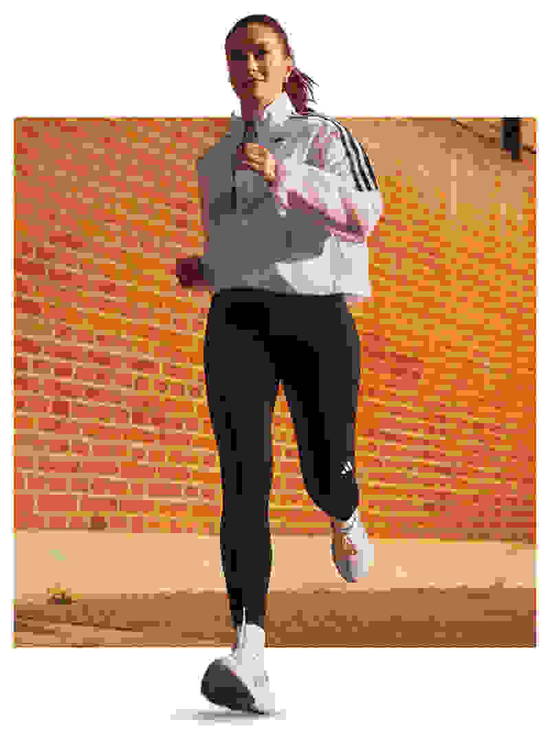 A young woman in athletic wear is standing in front of a brick wall, appearing to be in motion or preparing to run.
