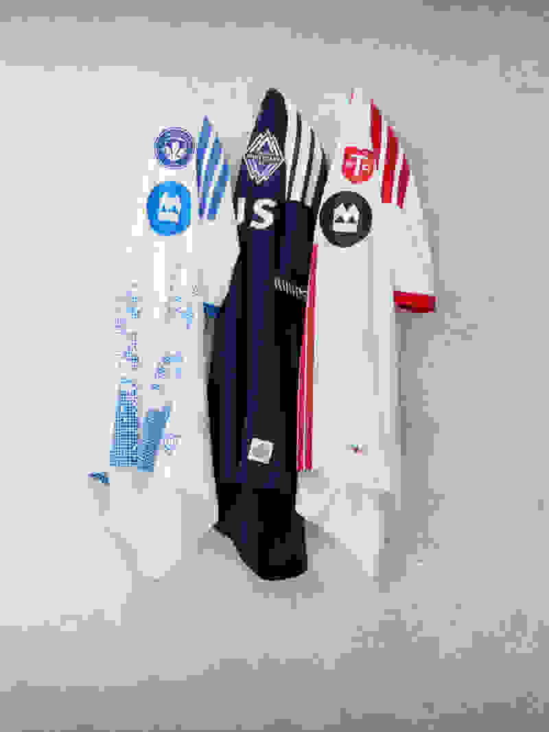 The image shows several soccer jerseys or uniforms hanging on a white wall, displaying various team logos and designs.