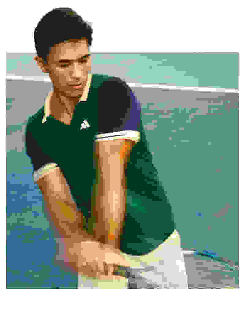 A young man in a green sports jersey is holding a tennis racket on a tennis court with a blue background.