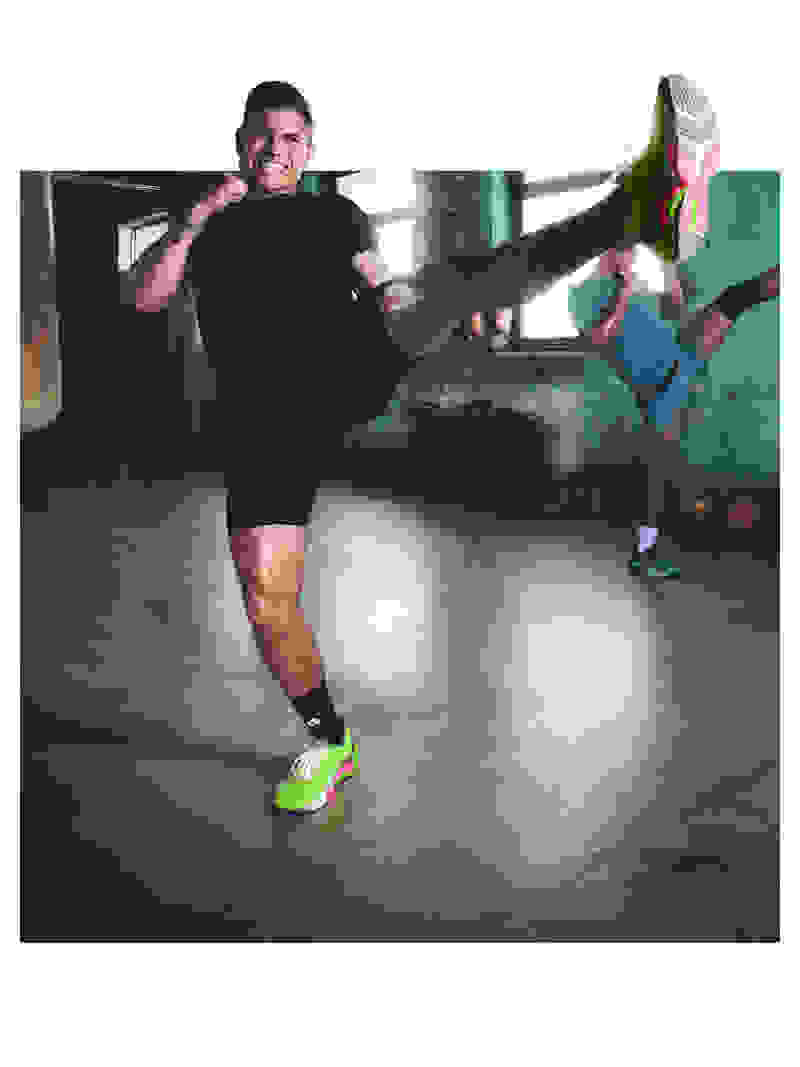 A man in black clothing and bright green sneakers is performing a martial arts kick in a dimly lit indoor setting.