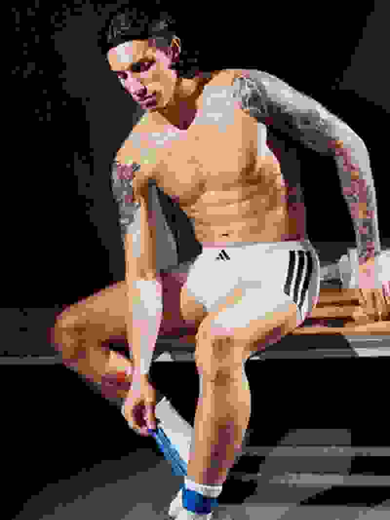 A shirtless, tattooed man with muscular physique sits on a wooden bench in a dimly lit setting.