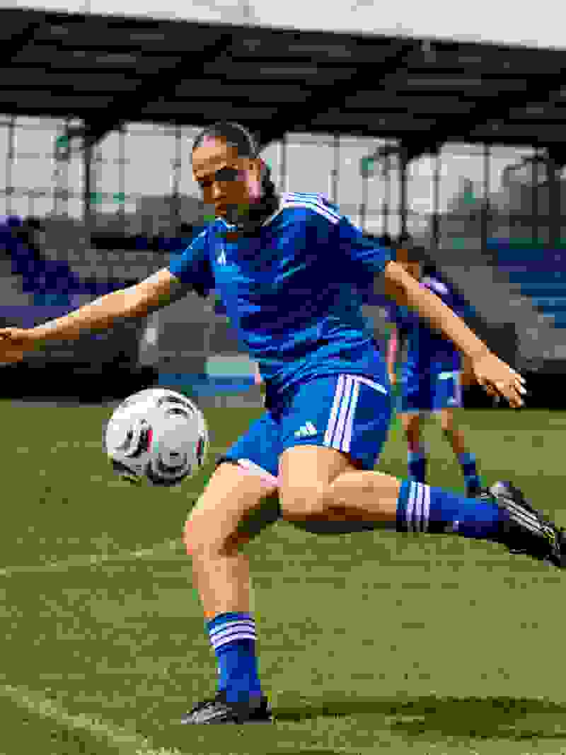 A soccer player in a blue uniform is running on a grassy field, with a stadium-like structure visible in the background.