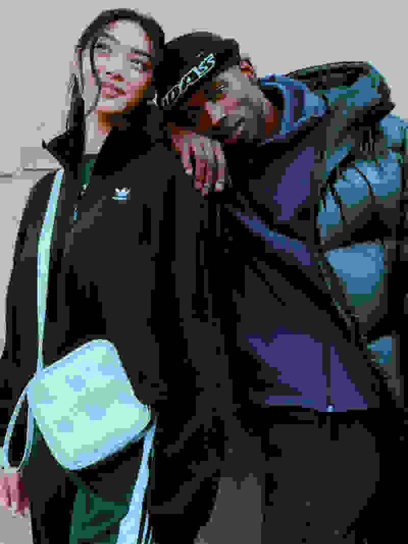 Two young people, a man and a woman, are embracing each other in a casual, affectionate manner. They appear to be dressed in casual, sporty attire, with the man wearing a black jacket and the woman wearing a black hoodie. The background suggests an indoor setting, possibly a room or a building.