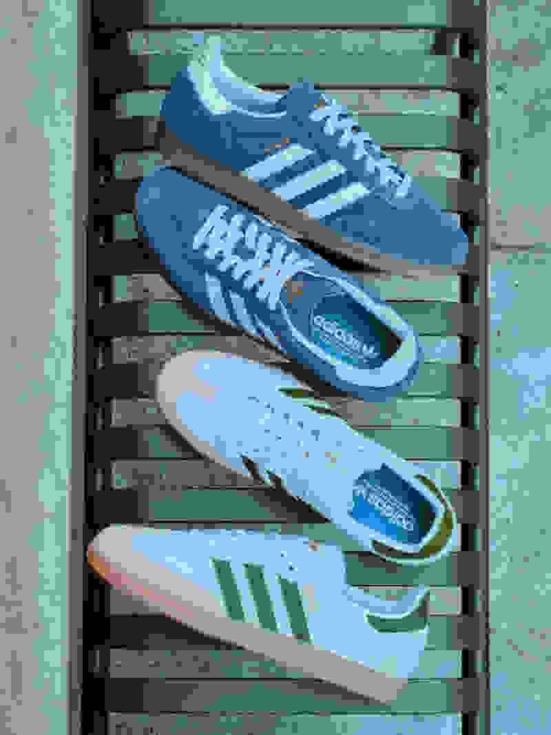 The image shows several pairs of Adidas sneakers in various shades of blue, white, and green, arranged on a wooden slat background.