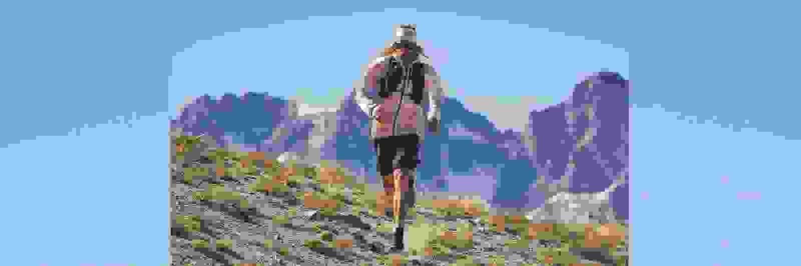 Eine Person mit rosa Mütze und Jacke steht auf felsigem, bergigem Terrain mit schneebedeckten Gipfeln im Hintergrund.