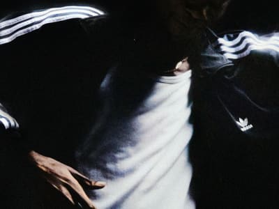 Person wearing a white tank top and a dark adidas Firebird jacket with the logo visible under dim lighting.